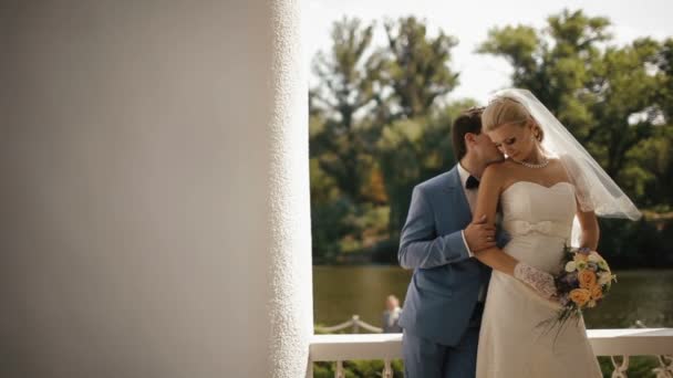 Romantic portrait of the charming couple of newlyweds. Handsome groom is tenderly kissing his bride in neck and face near the river. — Stock Video