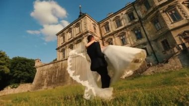 Yakışıklı damat günbatımı sırasında uzun şık gelinlik antik kalenin arka plan, onun sevimli gelin yuvarlak dönüyor.