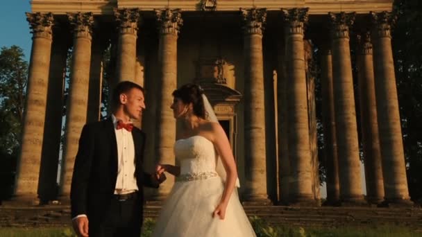 Tenderly kissing couple of attractive happy newlyweds over the background of old baroque style building during the sunset. — Stock Video