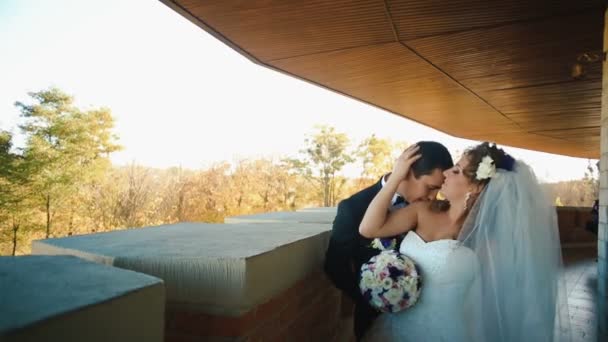 Όμορφος groom είναι φιλώντας γοητευτική κομψή νύφη του στο λαιμό και τον ώμο στην βεράντα. — Αρχείο Βίντεο
