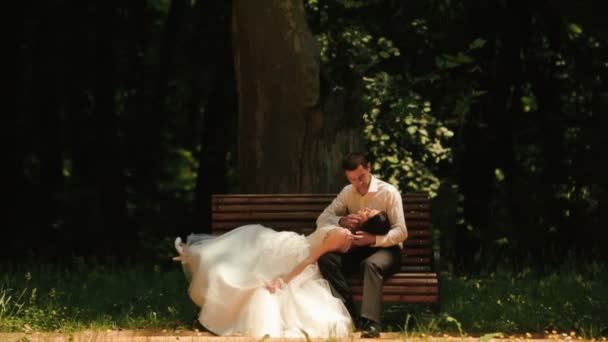 Romantische Atmosphäre. schöne Brautpaar küssen sich zärtlich. schöner Bräutigam streicht sanft über die Wange der Braut, während er auf der Bank im Park sitzt. — Stockvideo