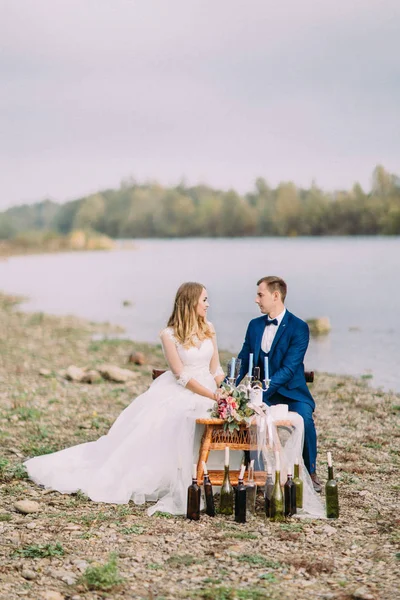 Os recém-casados felizes estão de mãos dadas enquanto se sentam na mesa de casamento na costa. Foto vertical . — Fotografia de Stock