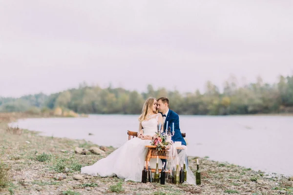 Gevoelige foto van de gelukkige knuffelen pasgetrouwden zitten aan de tafel van de bruiloft aan de kust. — Stockfoto