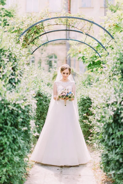 Красивая невеста смотрит на букет, стоя над аркой . — стоковое фото