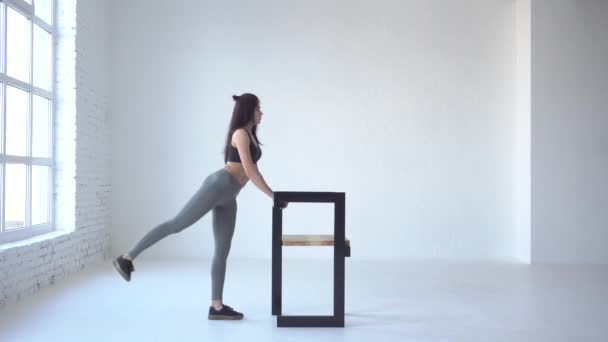 Sport oefeningen voor beginners. De jonge sporswoman doet de squat kickback terwijl leunend op de stoel in witte studio. 4 k-beeldmateriaal. — Stockvideo