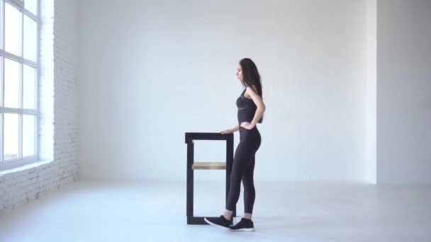 スポーティな細い女性のトレーナーは、白い分離スタジオの椅子にもたれながらキックをやっています。フルレングス サイドビュー。4 k. — ストック動画