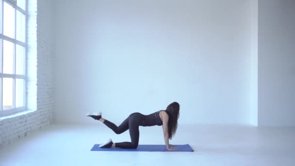 Fullängds vy av mager athletic trainer utför övning åsna kick på yogamattan i vita studio. 4 k film. — Stockvideo