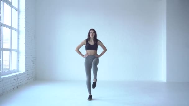 Aerobic für Anfänger. Sportkonzept. Die sportliche Trainerin im schwarzen Gymnastikanzug beugt die Beine im Knie und hebt sie im weißen Studio in die Höhe. 4k Filmmaterial. — Stockvideo