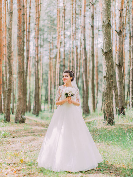 Full-length view of the charming bride with the wedding bouquet looking aside in the spring forest