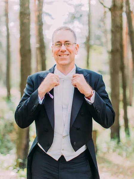 Half-length portrait of the cheerful groom corrects his white bow-tie. Forest location.