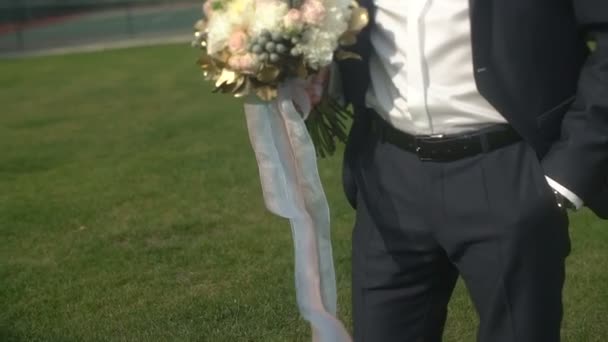 Caméra mobile de haut en bas. Vue d'ensemble du look du marié élégant en costume de luxe et lunettes de soleil tenant le bouquet de mariage et attendant sa mariée dans le jardin . — Video