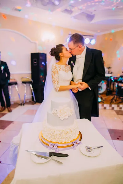 The white floral wedding cake at the background of the kissing newlywed couple.