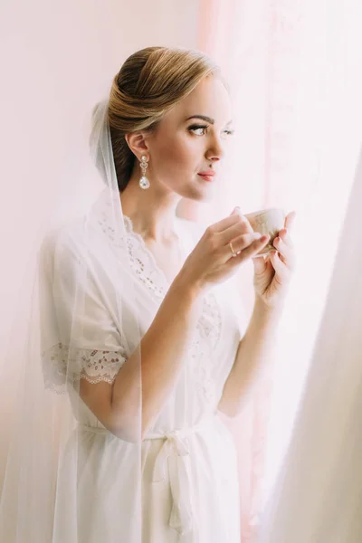 Beautiful bride holding the cup of tea while looking through the window. — Stock Photo, Image