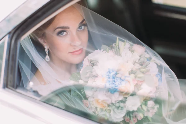 Vista horizontal de la novia bajo el velo que sostiene el ramo de bodas mientras está sentada en el coche . — Foto de Stock