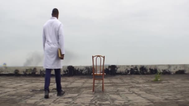 Pandangan belakang dari tidak diketahui african dokter duduk di kursi dengan buku. Dia sedang beristirahat di atap rumah sakit tua yang hancur . — Stok Video
