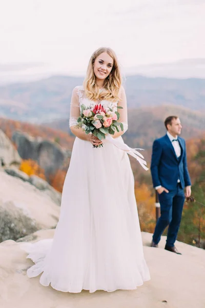 Belle mariée souriante sur le rocher au fond flou du marié . — Photo