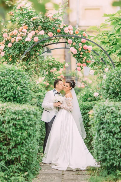 Groom está beijando a noiva encantadora na bochecha sob o arco de rosa . — Fotografia de Stock