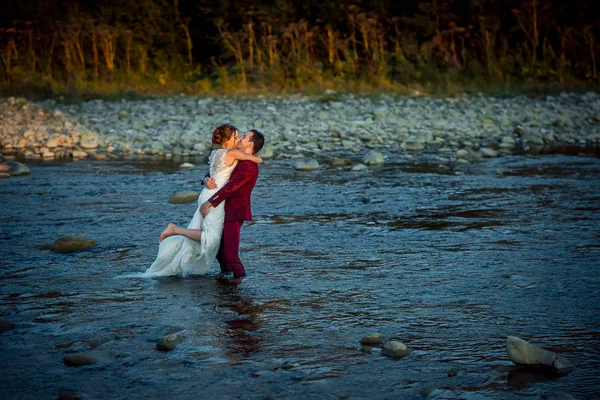 Boda horizontal de los hermosos recién casados. El novio en traje rojo está besando y levantando a su encantadora novia mientras está de pie en el medio del río durante la puesta del sol . — Foto de Stock