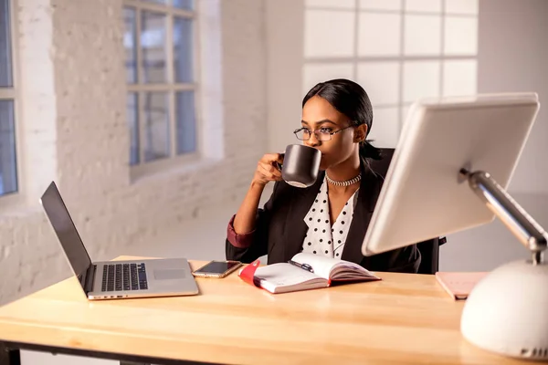 Empresária afro-americana sentada à mesa em seu escritório e bebendo café ou chá . — Fotografia de Stock