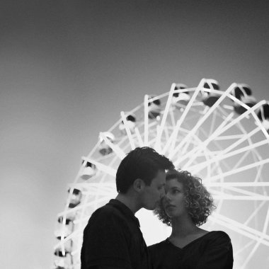 Dönme dolap içinde eğlence parkı önünde romantik genç çift. Kıvırcık kız ve erkek şefkatle kucaklamak