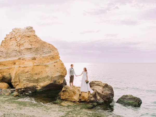 La vista orizzontale degli sposi felici che si tengono per mano mentre si trovano sulla scogliera tra l'oceano . — Foto Stock