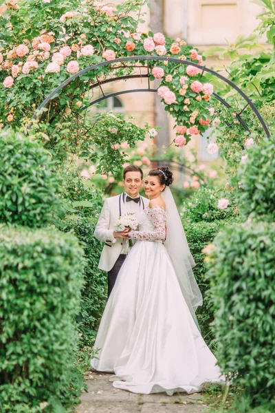 Foto completa dos recém-casados abraçando sob o arco de rosas . — Fotografia de Stock