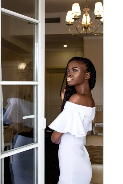 Georgeous junge afrikanisch-amerikanische Frau steht in der Nähe des großen französischen Fensters und posiert für die Kamera in einem schönen weißen Kleid. — Stockfoto