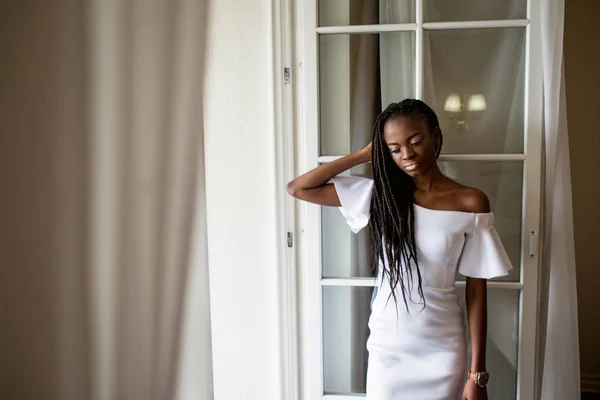 Bella ragazza afroamericana con dreadlocks o trecce africane in lungo vestito bianco in piedi vicino alla finestra . — Foto Stock