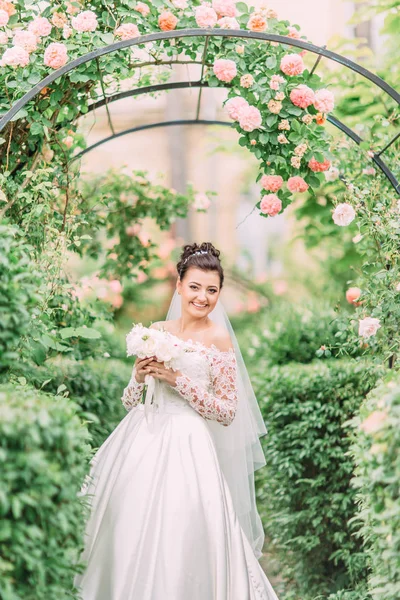 Retrato de meio comprimento da noiva feliz com buquê de casamento sob o arco com rosas . — Fotografia de Stock