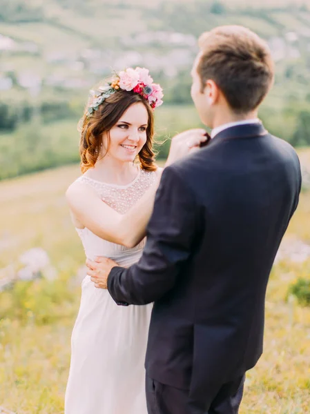 The smiling bride is correcting the bow-tie of the groom in the mountains.