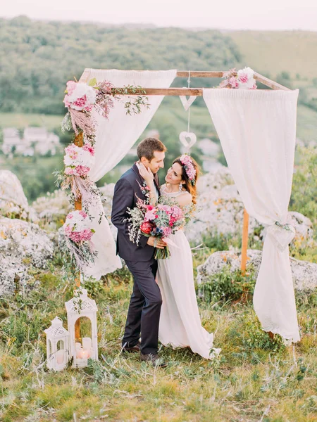 Повнометражний вигляд нареченої, що погладжує обличчя нареченого біля весільної арки в горах . — стокове фото