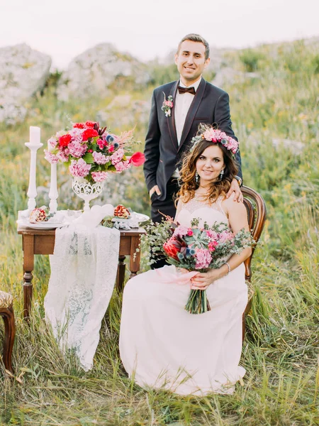 La novia con el ramo de bodas está sentada en la silla mientras el novio está de pie detrás de ella en las montañas . — Foto de Stock