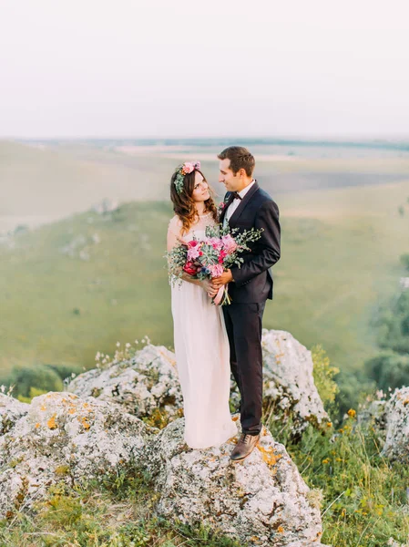 Nygift par tittar i ögonen på varandra stående på klippan vid bakgrunden av de gröna bergen. — Stockfoto