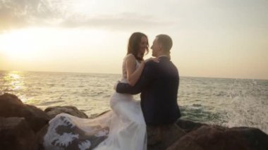 Mutlu büyüleyici yeni evli çift buzlu, şefkatle sarılma ve deniz geçmiş vasıl günbatımı sırasında konuşurken oturuyor.