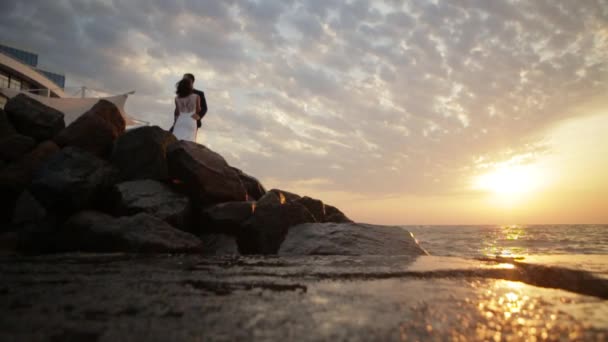 Rompiendo olas en las rocas durante la puesta del sol. Hermosa pareja de recién casados se abrazan tiernamente mientras están de pie en las montañas . — Vídeo de stock