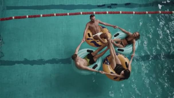 Vista superior a dos mujeres atractivas y dos chicos guapos divirtiéndose en los anillos de goma inflables en la piscina de hotel de lujo. 4k . — Vídeo de stock