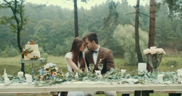 Przystojny mężczyzna w starodawny szmatką jest delikatnie całować jego kochanka w głowie podczas Data w Mglisty las. Siedzą przy stoliku pełen świece, liście, kwiaty i pyszne ciasto. 4k. — Wideo stockowe