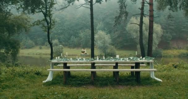Rörlig kamera mot bordet serveras för datum och dekorerad med löv, blommor, tvådelad tårta och ljus. Vackra naturen i dimmiga skogen och floden. 4k. — Stockvideo