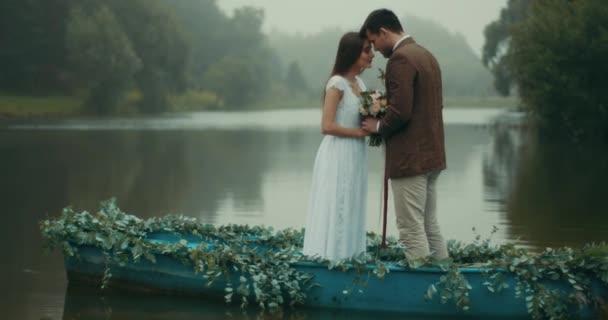 Hermosa pareja amorosa en tela vintage está sosteniendo el maravilloso ramo de flores y de pie cabeza a cabeza mientras flota en el romántico decorado con hierbas barco a lo largo del río brumoso. 4k . — Vídeos de Stock