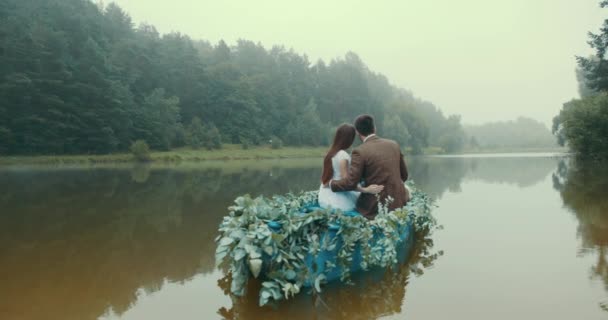 Vista trasera de la elegante pareja en pereza vintage flotando en un romántico barco decorado con hierbas verdes a lo largo del río brumoso. La mujer pone su cabeza en el hombro de su amante. 4k . — Vídeos de Stock