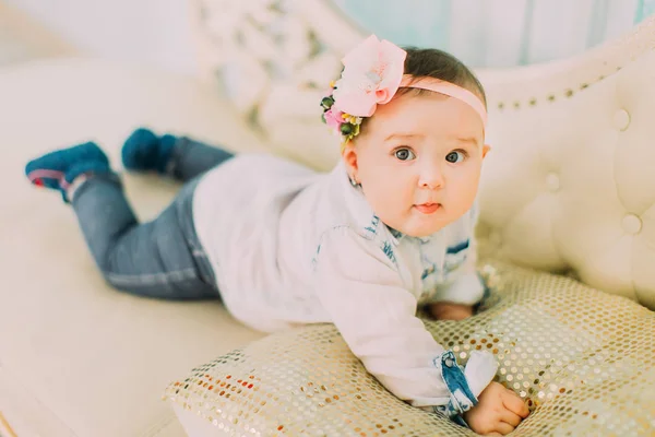 Pequeno bebê com flores e arco na cabeça está olhando para o lado esquerdo, enquanto deitado no sofá branco . — Fotografia de Stock