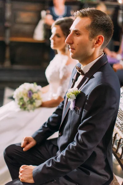O retrato lateral de meio comprimento do noivo sentado na cadeira durante a cerimônia de casamento na igreja no fundo borrado da noiva . — Fotografia de Stock