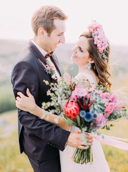 Zijaanzicht van de gelukkig knuffelen pasgetrouwden houden de enorme bruiloft boeket van exotische bloemen. — Stockfoto