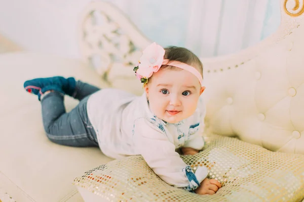 The front portrait of the little baby with the bow and flowers on the head lying on the white sofa. — Stock Photo, Image