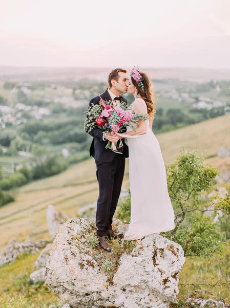 Preciosa foto de los recién casados besándose con el enorme ramo de bodas mientras están de pie sobre la piedra en las montañas . — Foto de Stock