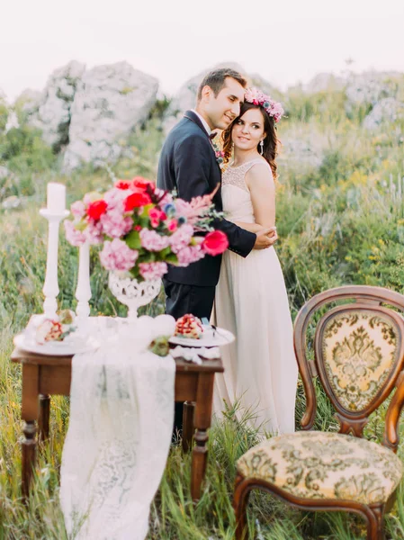 The hugging newlyweds behind the blurred composition of the wedding table set in the mountains. — Stock Photo, Image
