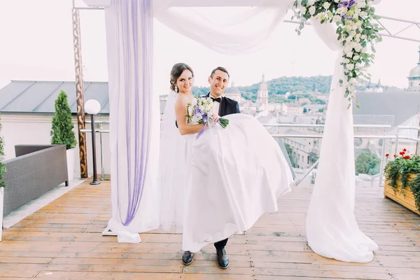 Feliz noivo está carregando a noiva sorridente perto do arco de casamento . — Fotografia de Stock