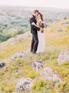 Dağlardaki ayakta sarılma yeni evliler tam uzunlukta görünümünü. Damat buket tutuyor.