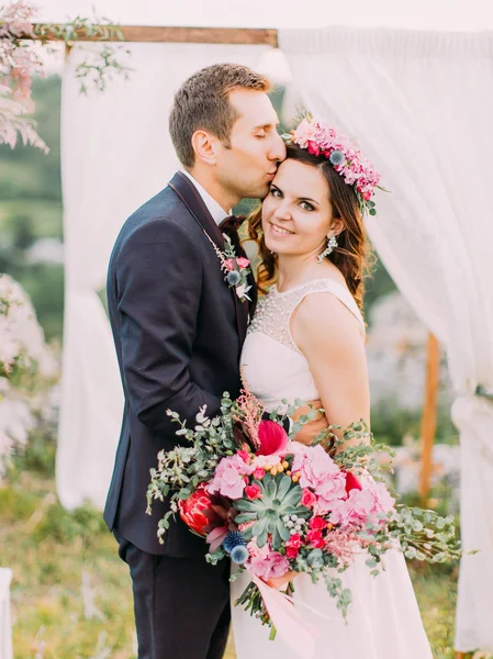 Close-up outdoor portrait of the cheerful groom kissing the smiling bride in the forehead near the arch.