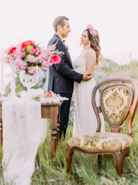 The vertical view of the hugging newlyweds behind the blurred wedding table set. — Stock Photo, Image